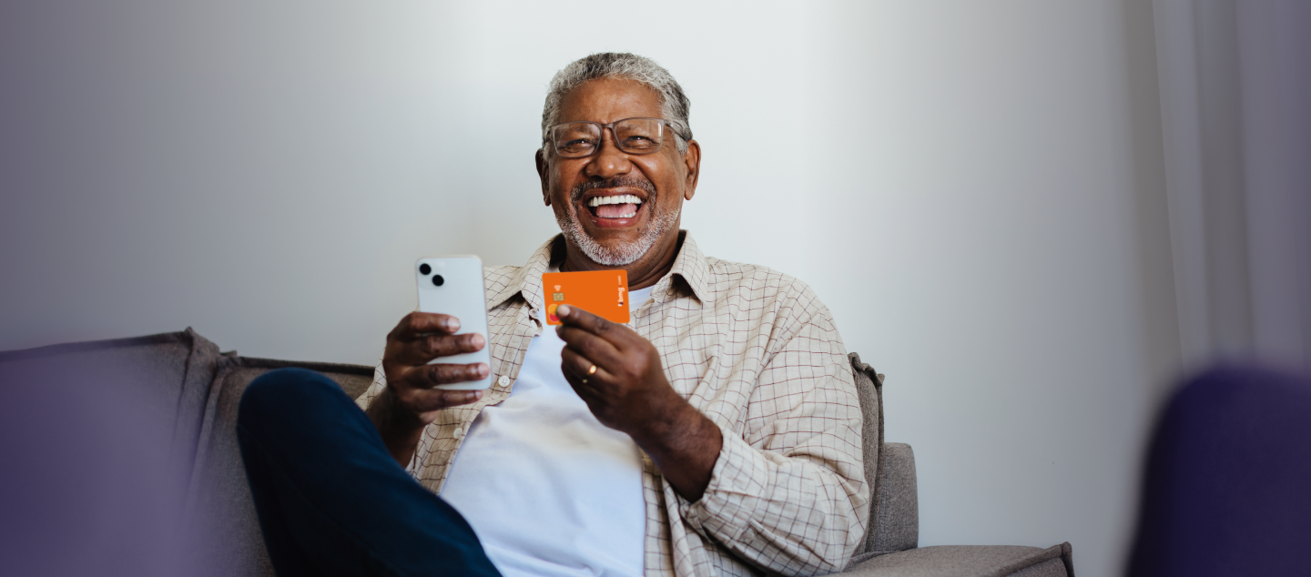 Imagem de um homem idoso preto, sentado no sofá, enquanto segura um celular em um mão e um cartão laranja na outra mão.
